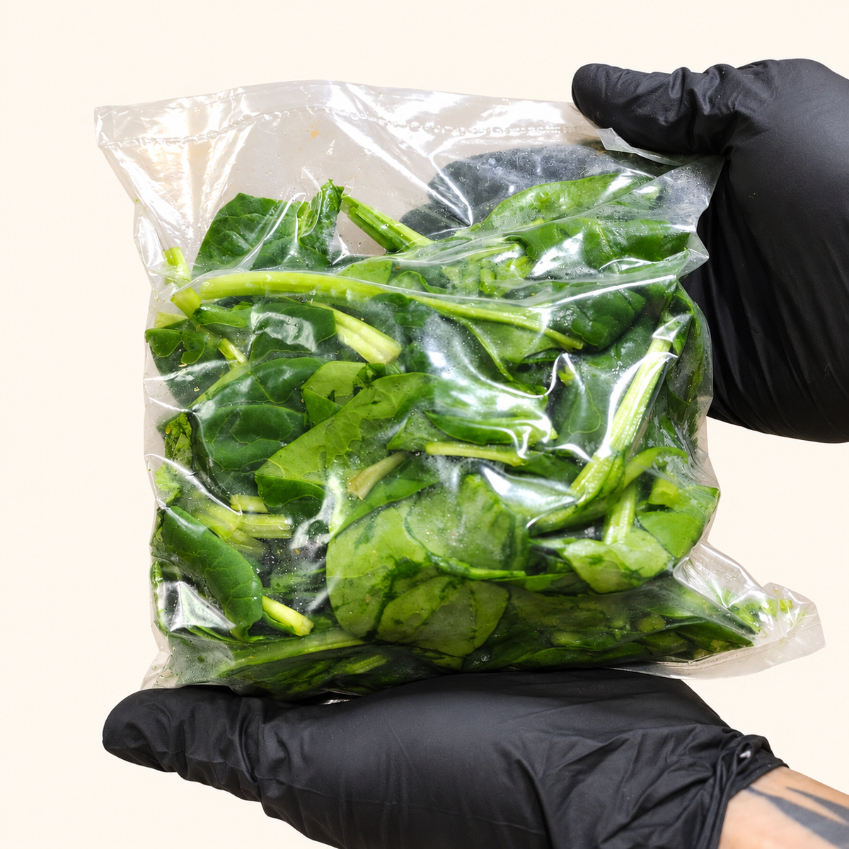 Bolsa de espinaca fresca de 300 g sostenida con ambas manos enguantadas, sobre fondo crema.