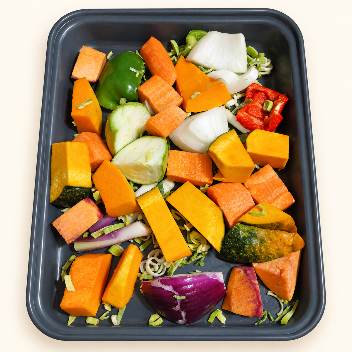 Detalle de vegetales crudos para horno, cortados y dispuestos en una bandeja oscura sobre fondo crema.